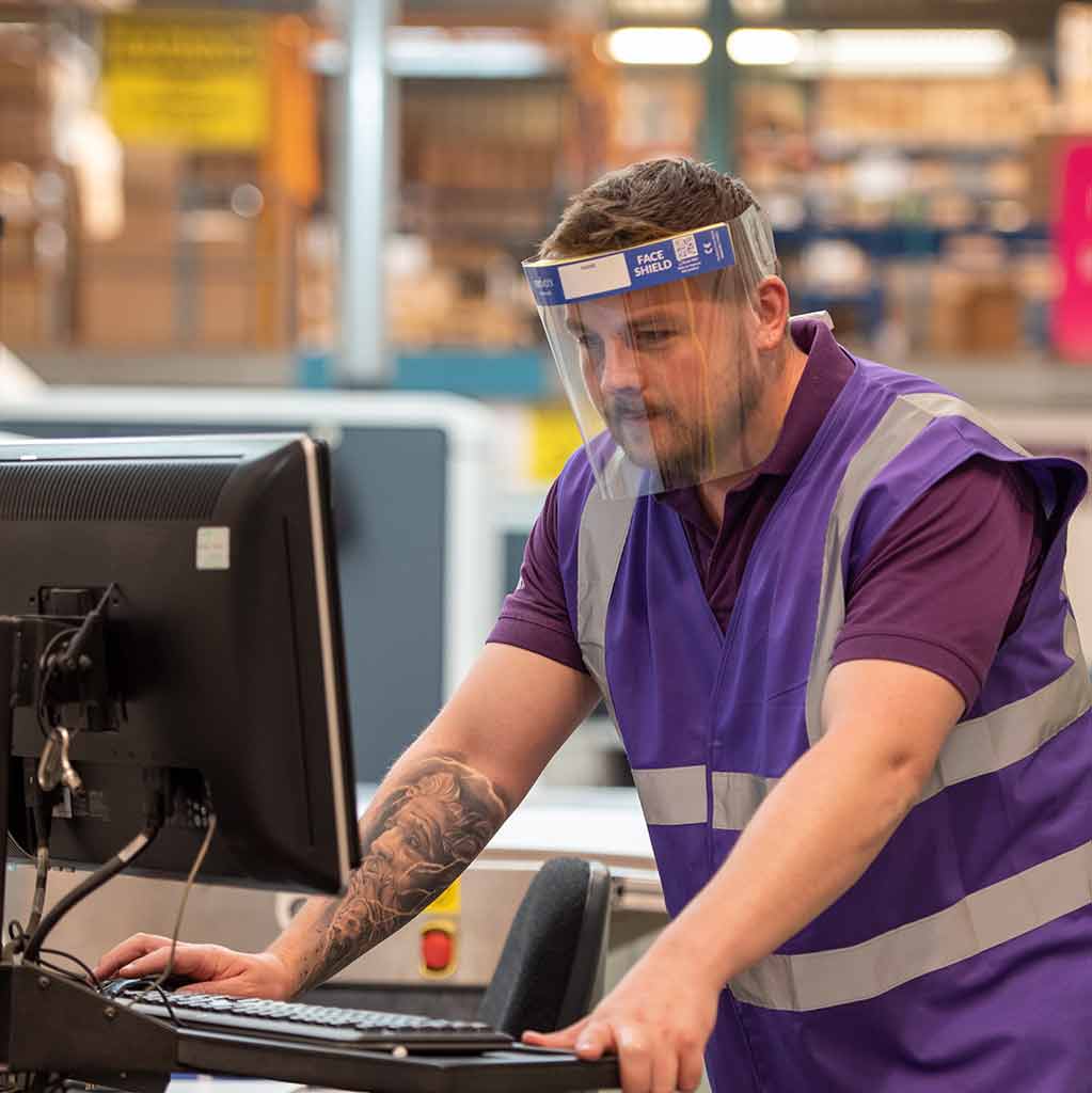 Man with a tattoo on arm working on a computer in a warehouse wearing a purple hi vis jacket and Medworx face shield.